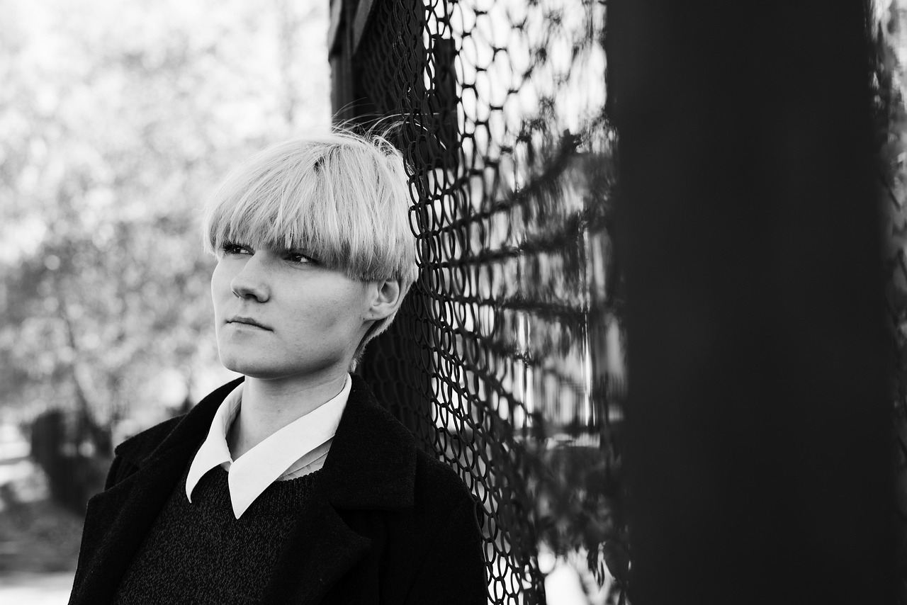 Čiernobiela fotografia osoby v čiernom kabáte a bielou košeľou, opierajúca sa o kovovú sieť.