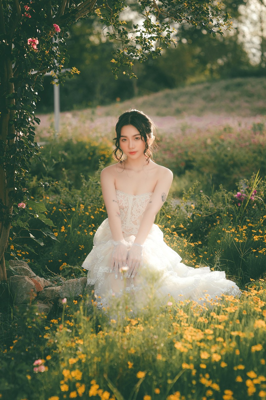 Žena v bielych šatách s kvetinovými prvkami sedí v parku plnom kvetov.